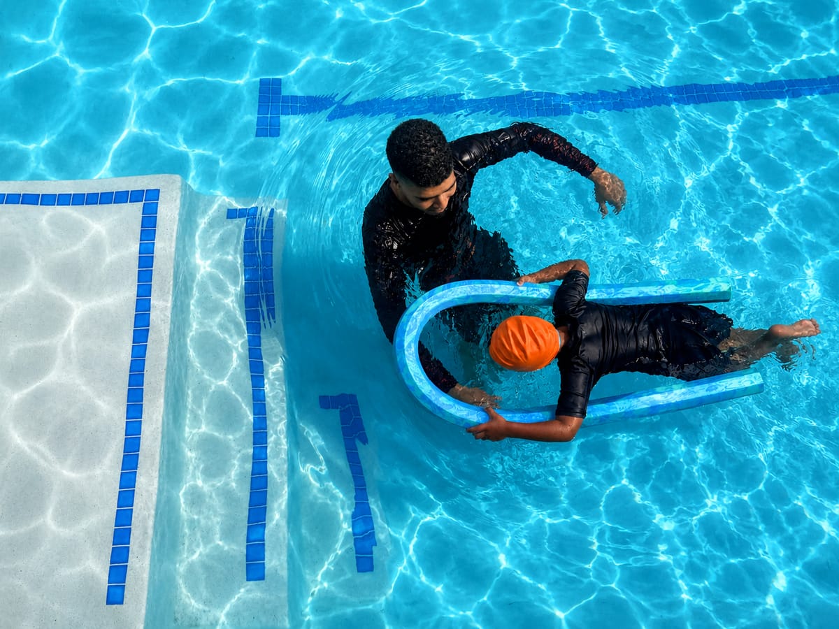 Overhead view of an instructor guiding a child with a pool noodle beside the pool steps