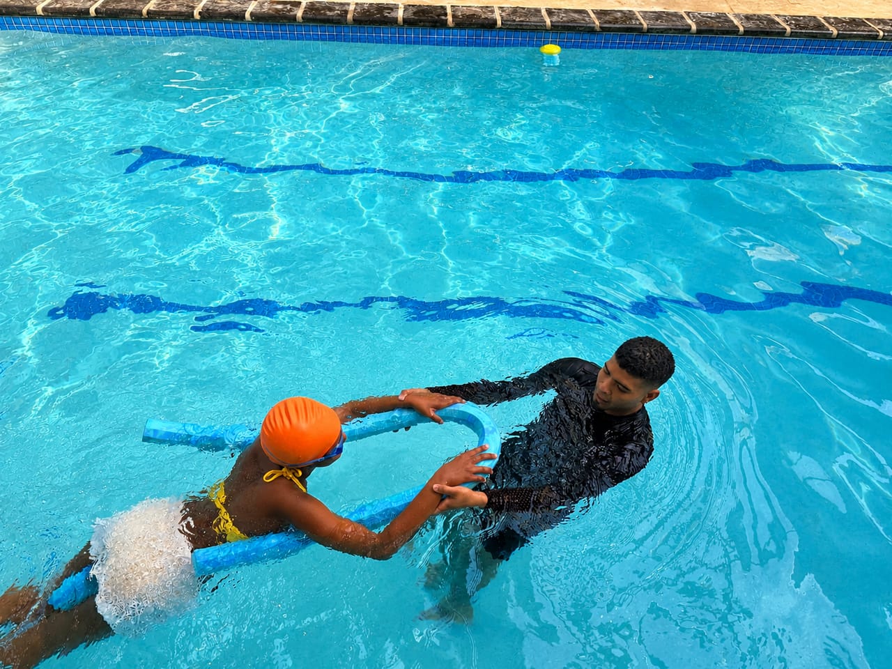 Instructor guiding a child along a pool noodle, encouraging them to reach forward