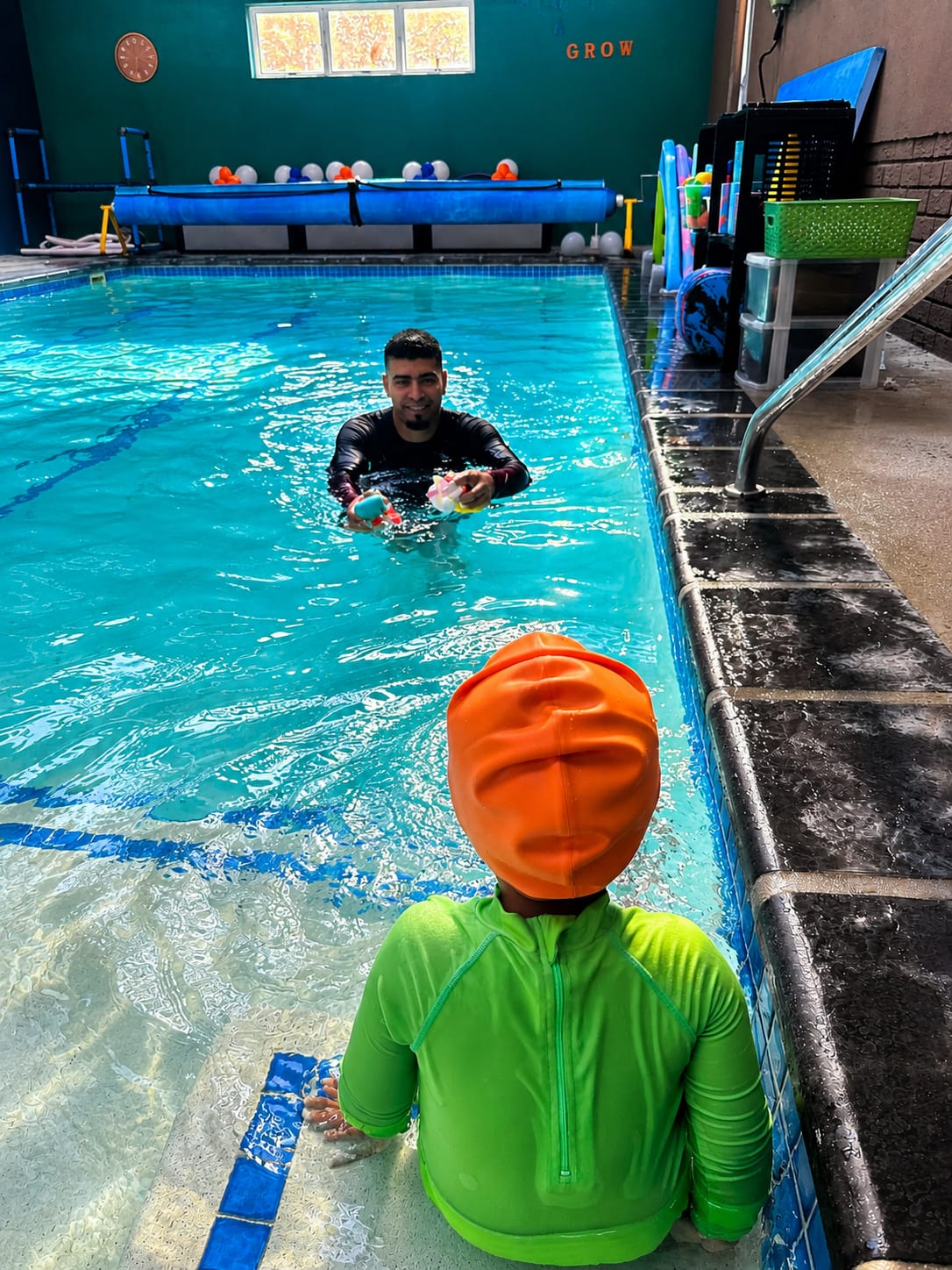 A child sitting at the poolside while the instructor encourages them into the water