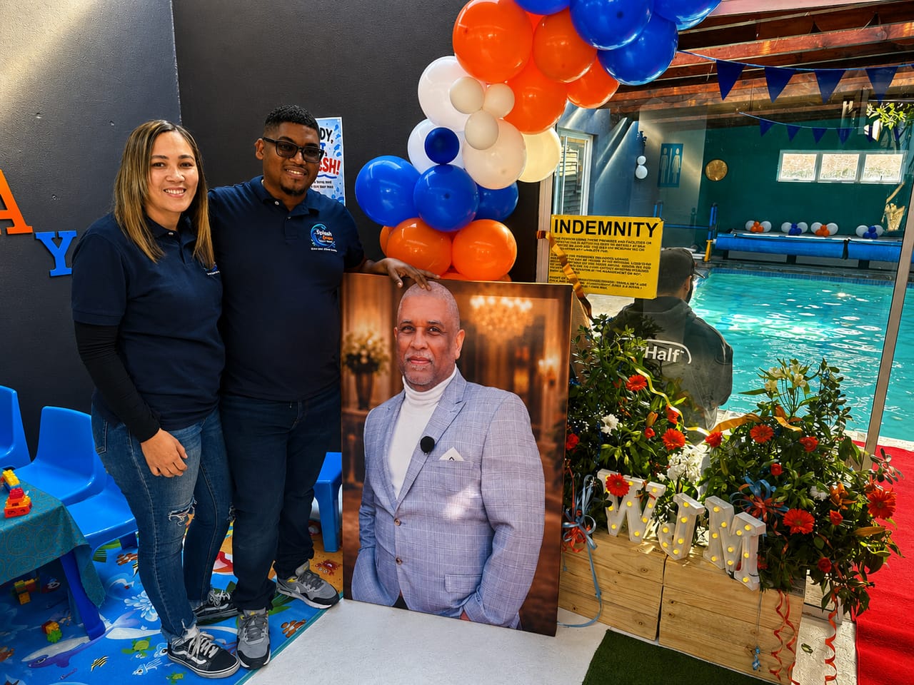 Two Splash & Grow team members beside a framed portrait and floral display at the open day, with the indoor pool behind