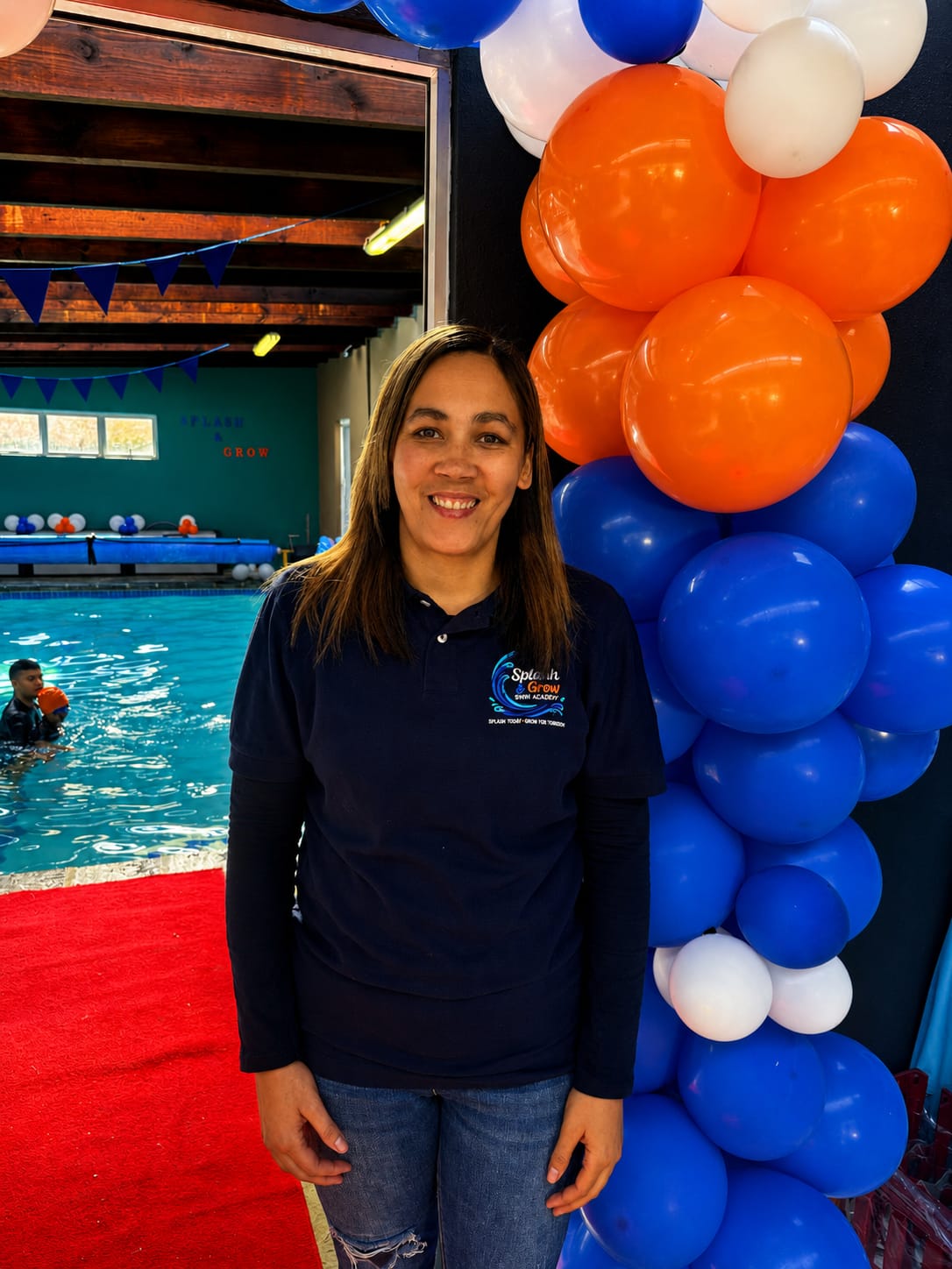 A smiling Splash & Grow instructor beside the heated pool, framed by open day balloons, as a child swims